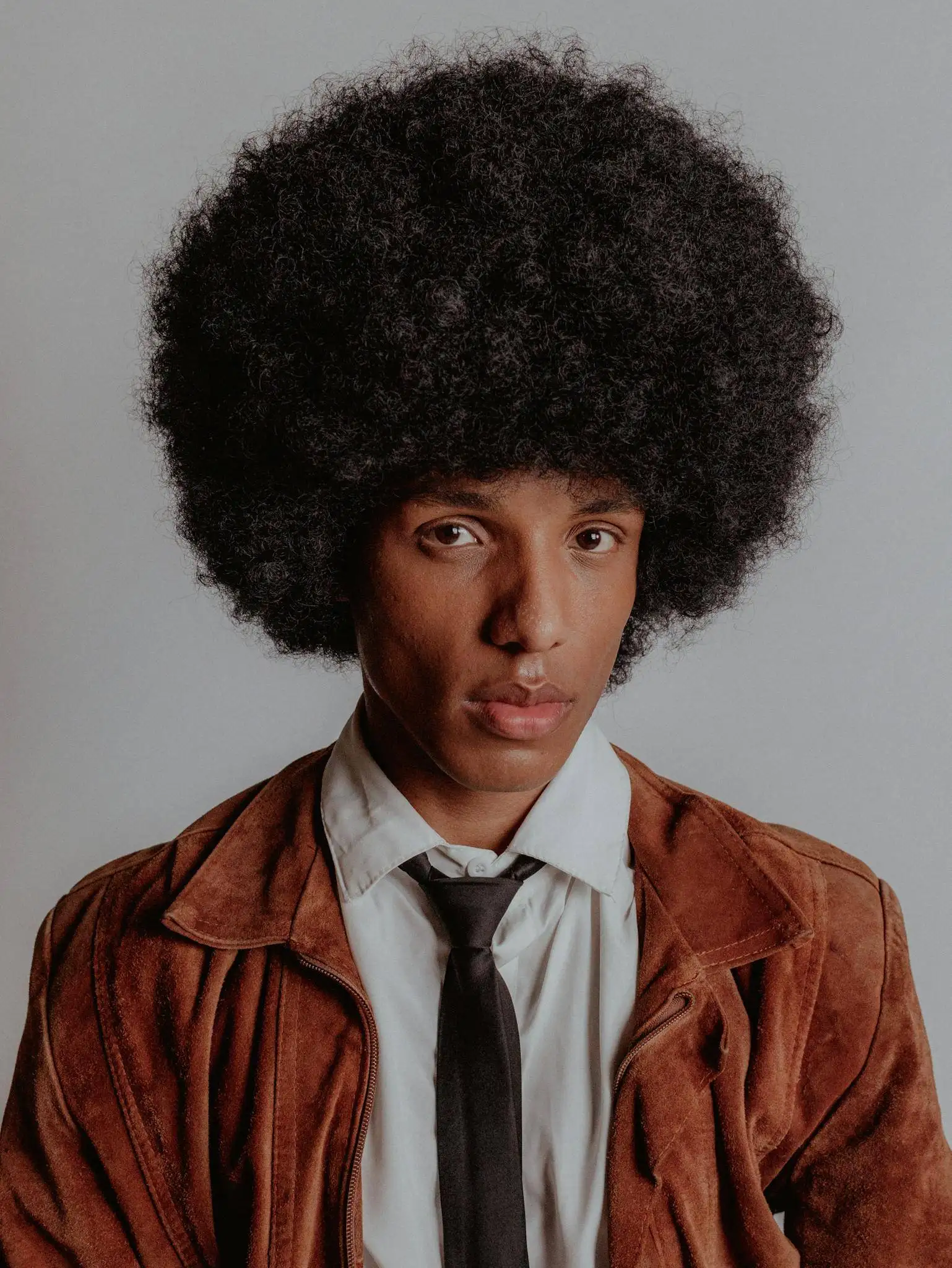Home 6 Young man in brown jacket with an afro hairstyle poses against a neutral background.